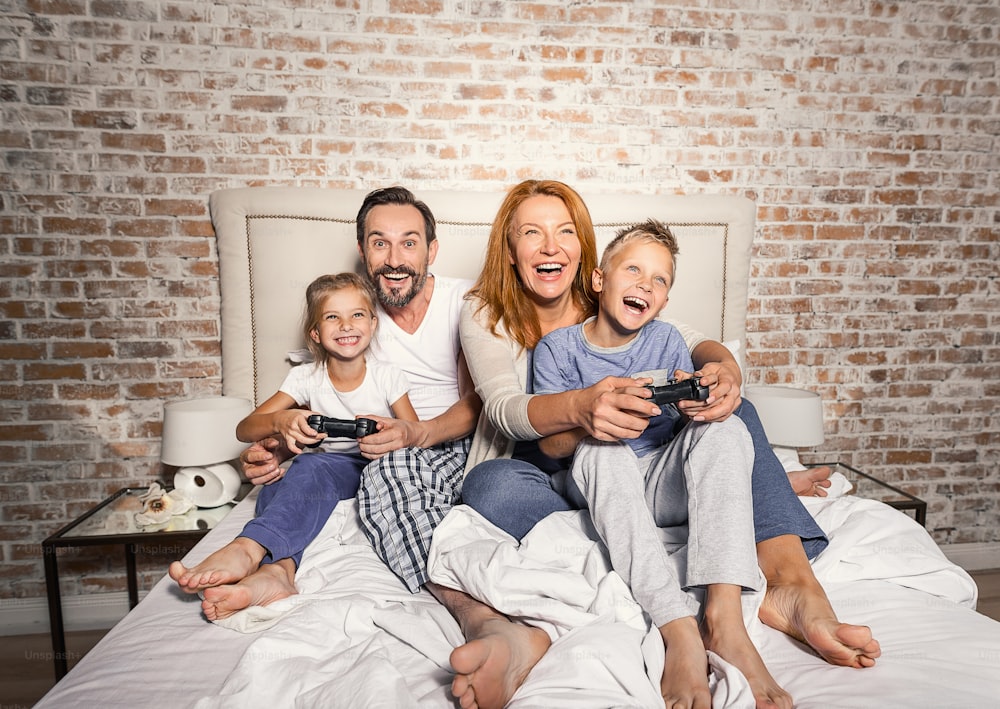 Fun times together. Mature smiling parents playing on console with their children enthusiastically while sitting on bed at home