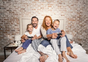 Fun times together. Mature smiling parents playing on console with their children enthusiastically while sitting on bed at home