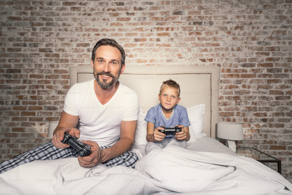 Padre feliz e hijo sonriente jugando videojuegos en la cama mientras sostienen joysticks en sus manos