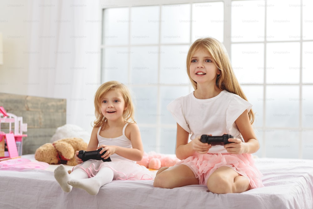 Des enfants enthousiastes gardant des joysticks dans leurs mains tout en jouant à des jeux vidéo. Enfants assis sur un lit moelleux en face d’une large fenêtre. Les ours en peluche sont près d’eux