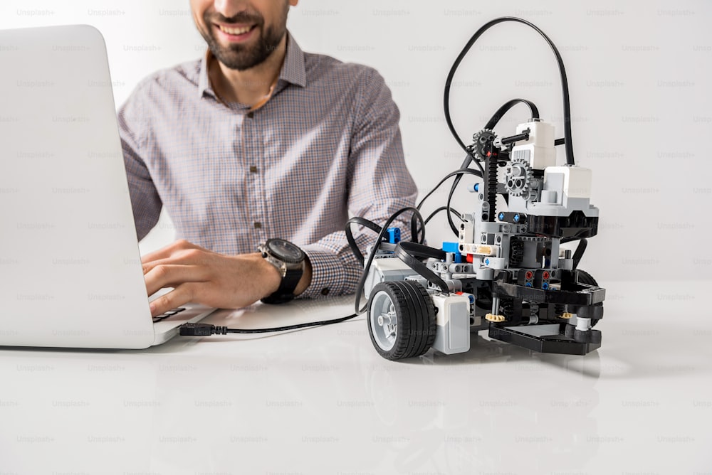 Enjoying modern technology. Cheerful professional man is sitting at table and working on gadget. Isolated. Focus on robotic toy