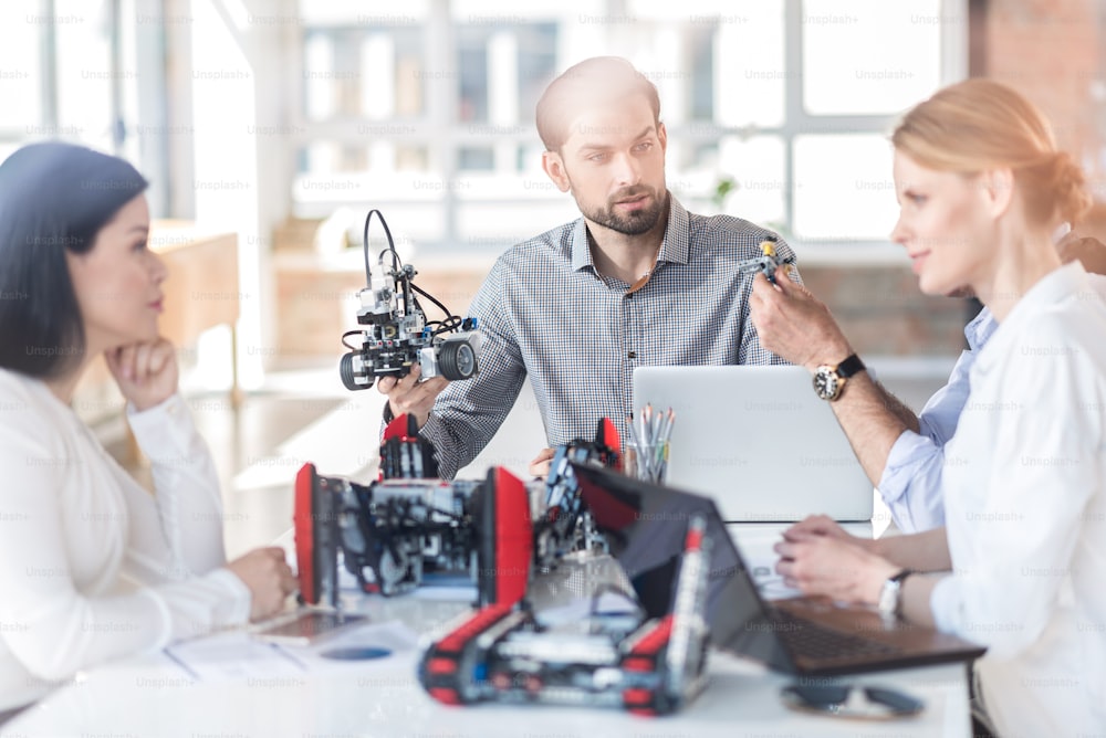 Robotic technologies in use. Positive professional engineers sitting at table and constructing robot. Selective focus on young bearded man holding device