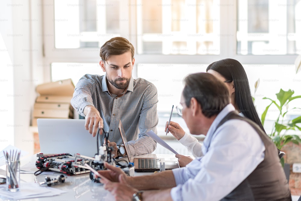 Tech progress. Selective focus on handsome bearded man is sitting with colleagues at table. He is pointing to robot with interest