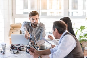 Tech progress. Selective focus on handsome bearded man is sitting with colleagues at table. He is pointing to robot with interest