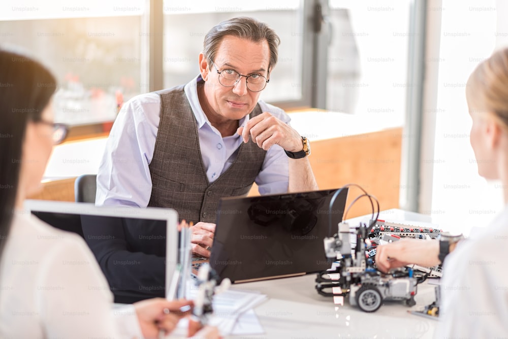 Looking for new ideas. Group of engineers sitting around table while working on laptop. Focus on portrait of serious senior man looking at camera thoughtfully