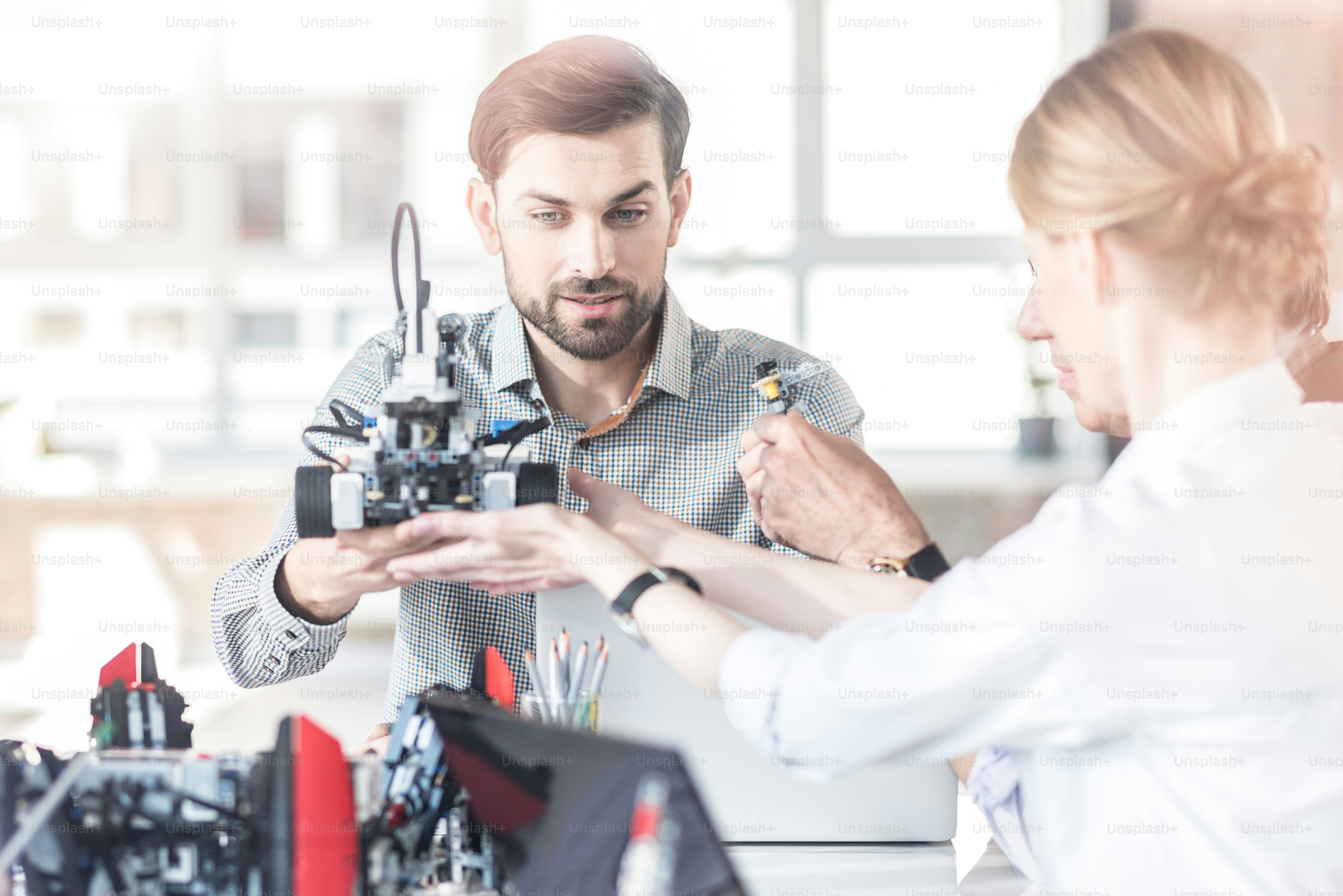 Verifizierung eines modernen Geräts. Selektiver Fokus auf einen entzückt lächelnden, selbstbewussten Kerl, der am Tisch sitzt und den Roboter mit seinem Kollegen hält. Blick von der Glaswand