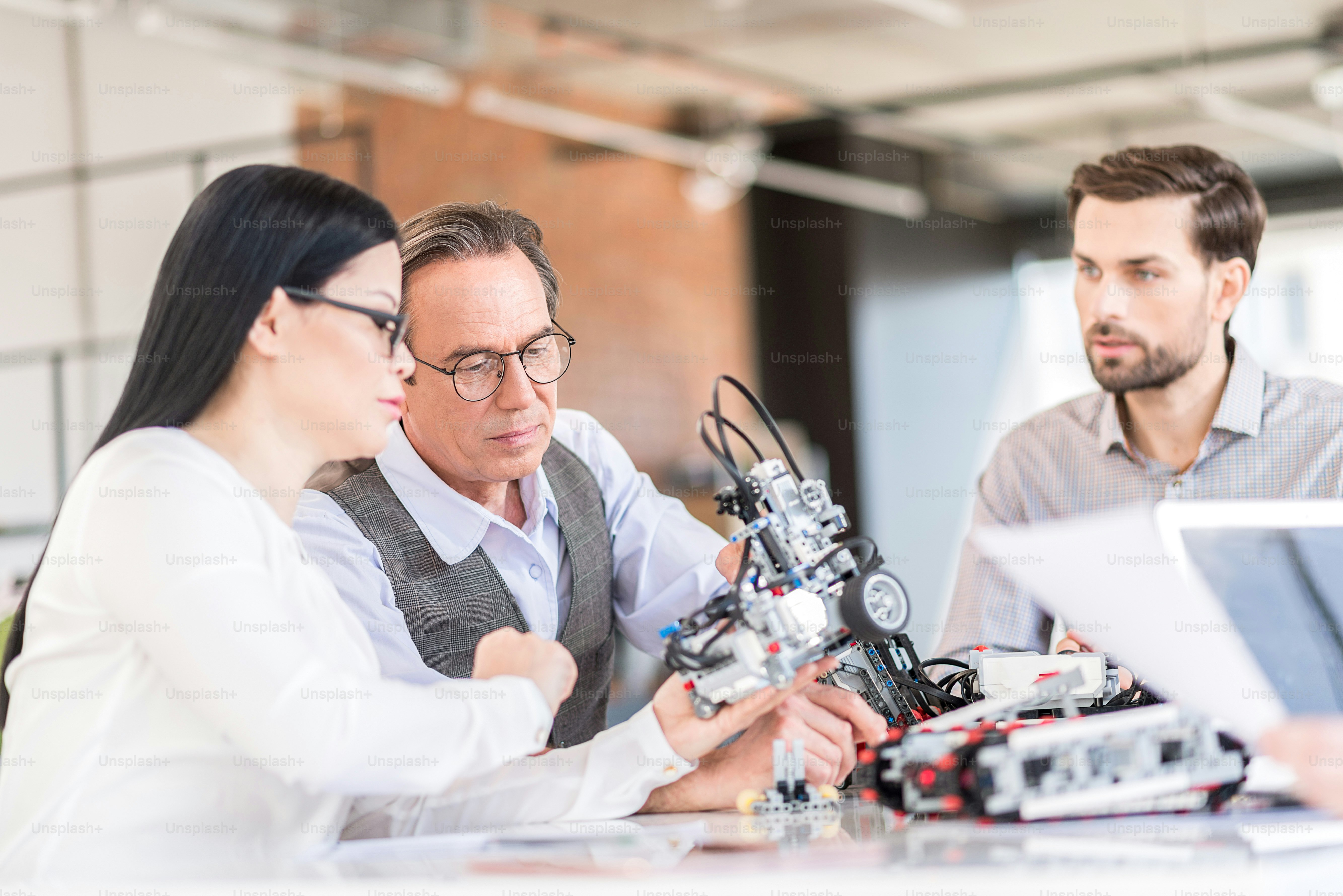 Notre projet technologique. Des travailleurs concentrés attentifs assis à table et tenant un robot tout en exprimant leur intérêt. Accent sélectif sur l’homme âgé en lunettes regardant attentivement le mécanisme