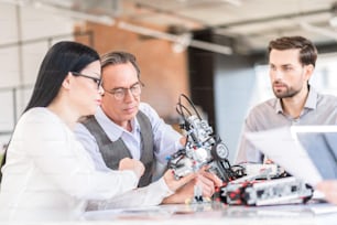 Our tech project. Attentive concentrated workers sitting at table and holding robot while expressing interest. Selective focus on senior man in glasses looking at mechanism intently
