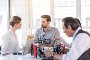 Creation of project. Group of inventors are developing robot construction. Young bearded man is demonstrating device while looking at woman intently