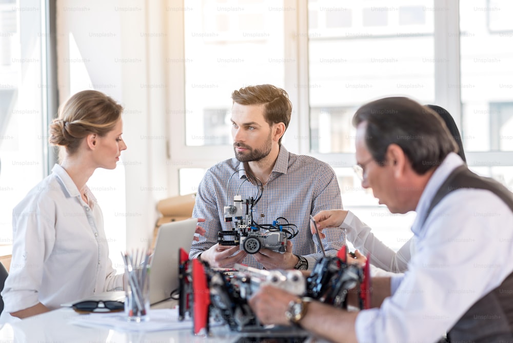 Creation of project. Group of inventors are developing robot construction. Young bearded man is demonstrating device while looking at woman intently