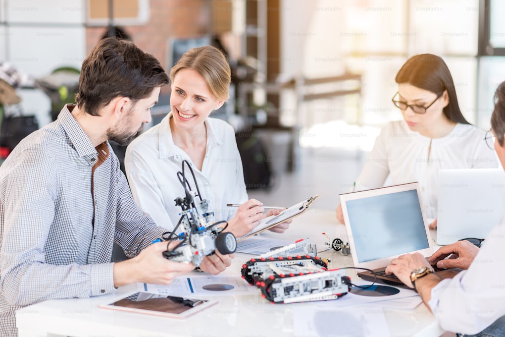 Much act to do. Selective focus on attractive professional lady is making notes while young bearded man is testing robot in office. Woman is sitting and looking at him with joy