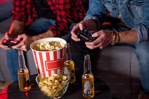 Playing together. Close-up of game console being in hands of young men. They are sitting in cozy apartment with bottles of beer, potato chips and popcorn