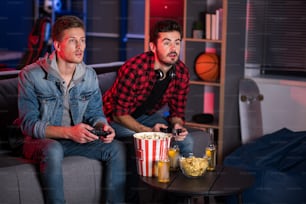 Weekend entertainment. Involved trendy cute guys are sitting on couch and holding joystick while playing home video games. They are looking at screen absorbedly. Copy space in the right side