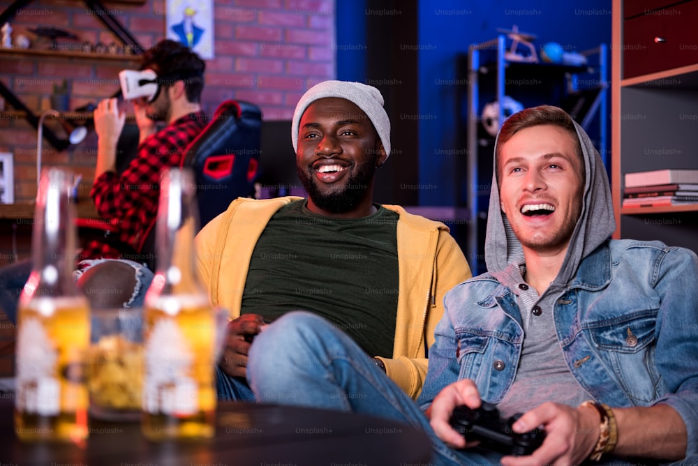 Adult toys. Joyful best international friends are using video game console and sitting on sofa while expressing excitement. Cute guy is wearing VR glasses in background. Bottles of beer in foreground