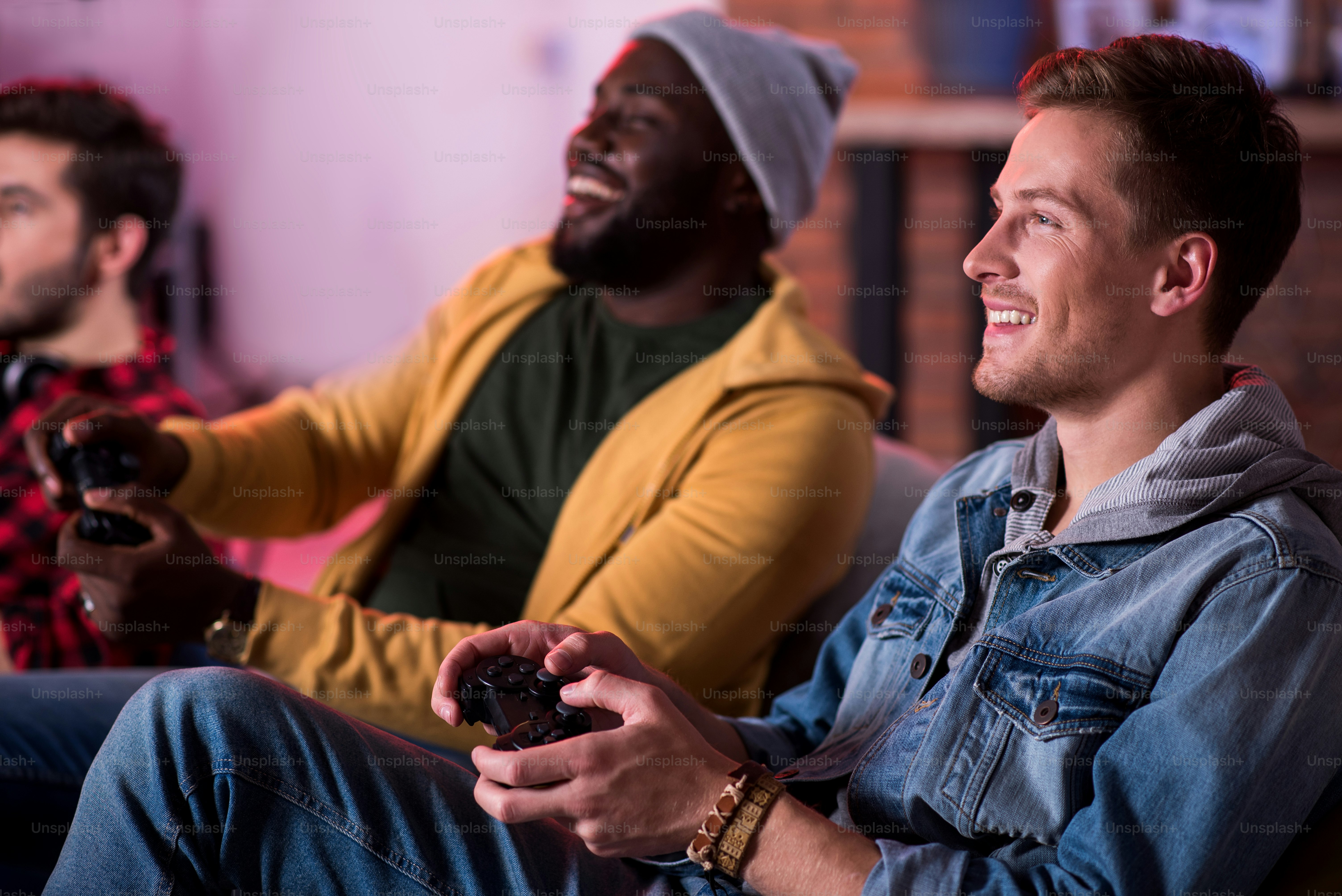 Fiesta de adultos. Los chicos atractivos y alegres de moda están disfrutando de la estación de juego. Están sentados en el sofá y expresando alegría mientras sostienen el joystick y miran el monitor. Enfoque selectivo