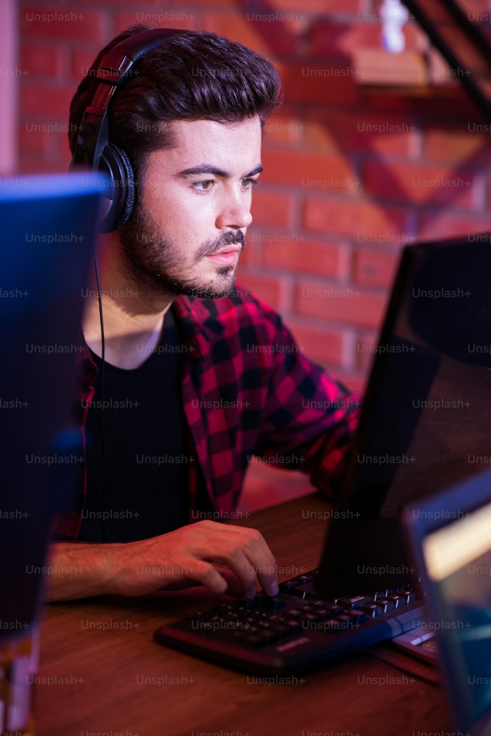 Important moment. Serious young stylish guy in headphones is sitting at desk and developing his strategy while playing video games on modern computer. He is looking at screen with concentration