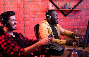 Selective focus of positive african man in headphones is sitting at table with friend and playing on computer. They are expressing gladness. Joyful bearded guy is holding steering wheel in foreground