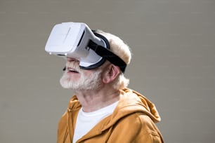 Enchanted male pensioner having fun while using virtual googles. Isolated on grey background