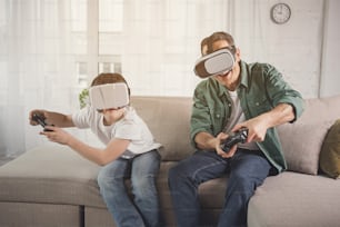 Aufgeregte Eltern und Kinder haben Spaß mit Joysticks und VR-Geräten. Sie sitzen auf der Couch und lächeln
