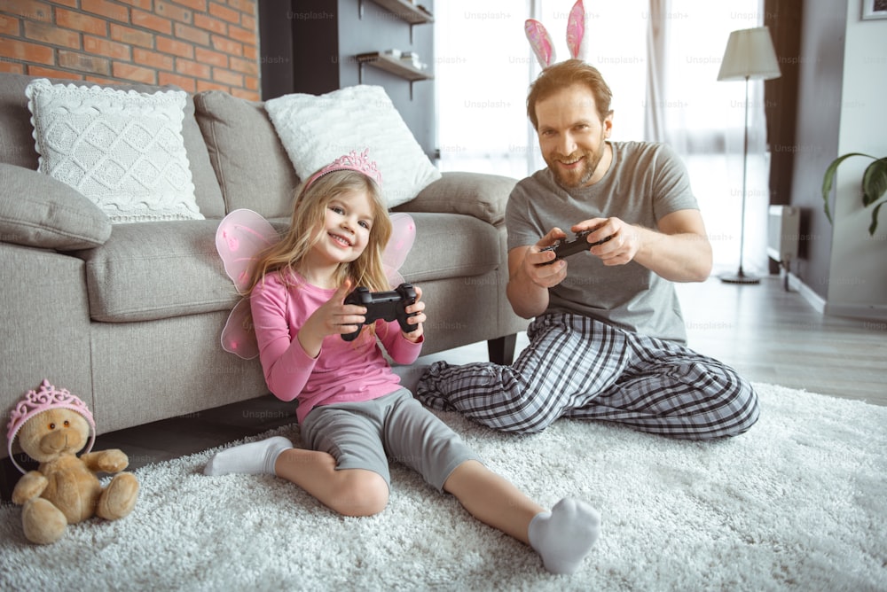 Family entertainment. Full length portrait of happy girl using joystick while looking forward with excitement. Her dad is sitting on floor and smiling