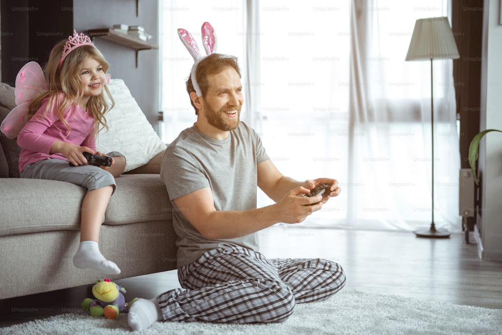 Excited father and daughter are playing video games together at home. They are relaxing at couch and laughing