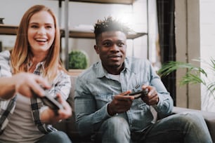 Portrait of cheerful pretty girl and handsome afro american guy sitting on couch and using joysticks. Focus on young man in denim shirt