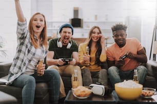 Young people having fun while sitting on couch. Guys using joysticks while girls holding bottles of beer and supporting gamers