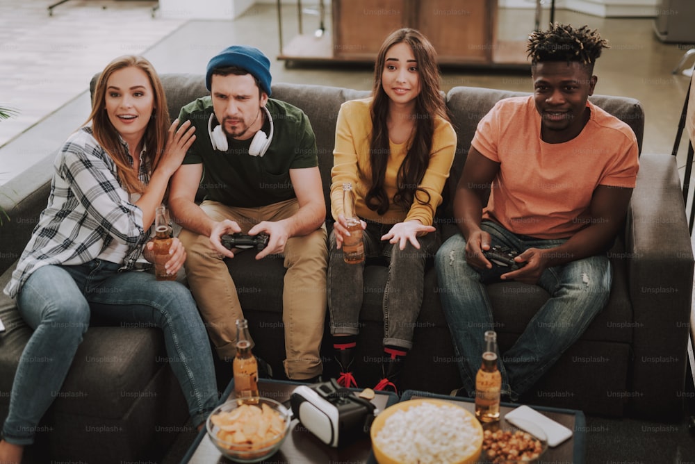 Portrait of young men using joysticks while ladies holding bottles of beer and smiling. Friends sitting on couch at home