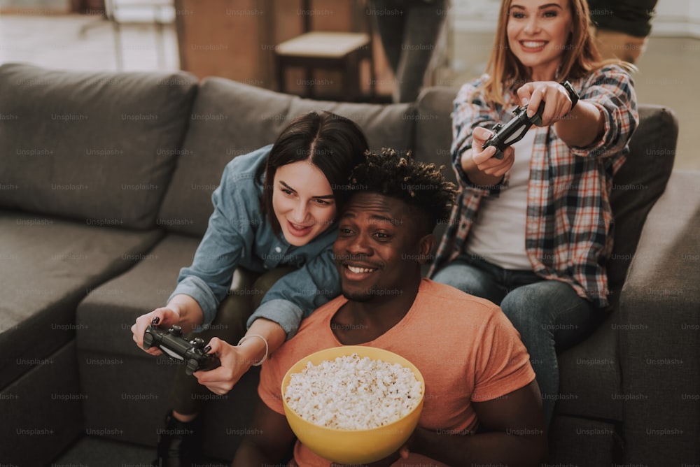 Beautiful young ladies sitting on couch and playing video games while afro american guy smiling and holding popcorn