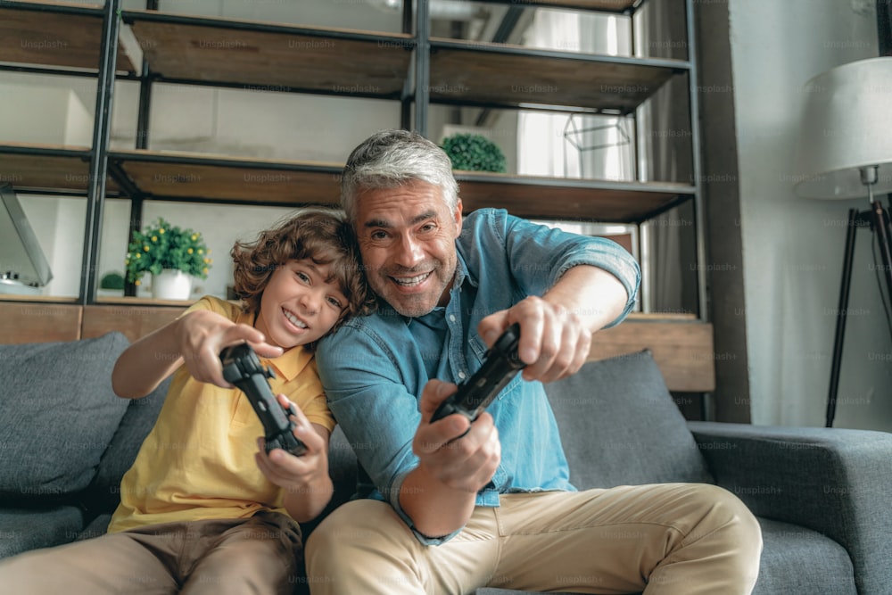 Happy mature father playing with cheerful son in video games, sitting on sofa