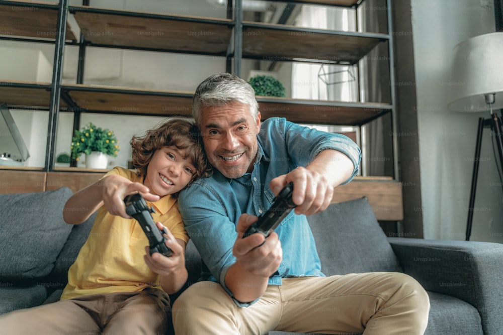 Happy mature father playing with cheerful son in video games, sitting on sofa