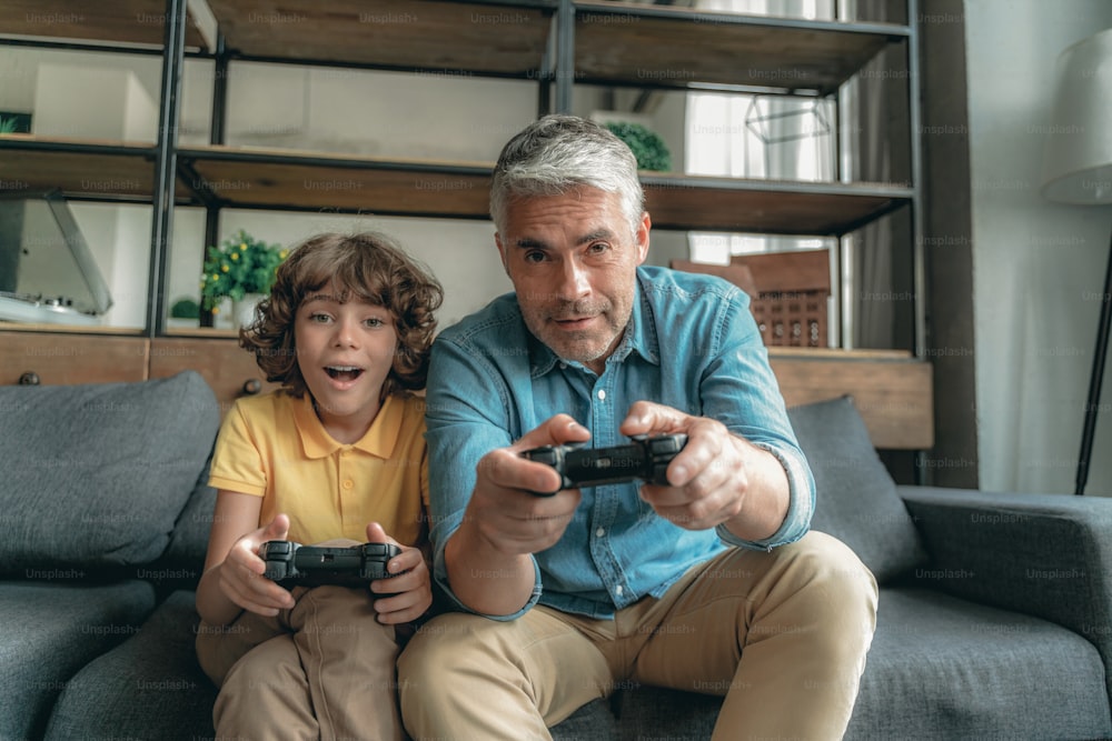 Excited son sitting near father on couch and playing in video games
