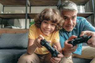행복하고 웃는 아버지는 어린 아들 근처에 앉아 비디오 게임을 하고 있다