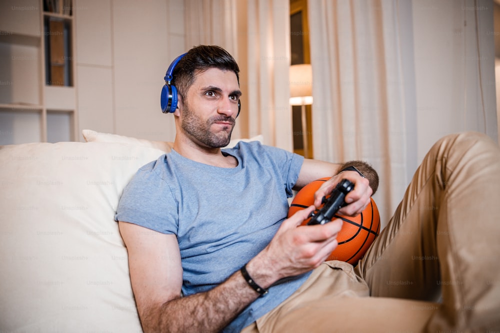 Young man in headphones is sitting on couch and entertaining himself with joypad and ball