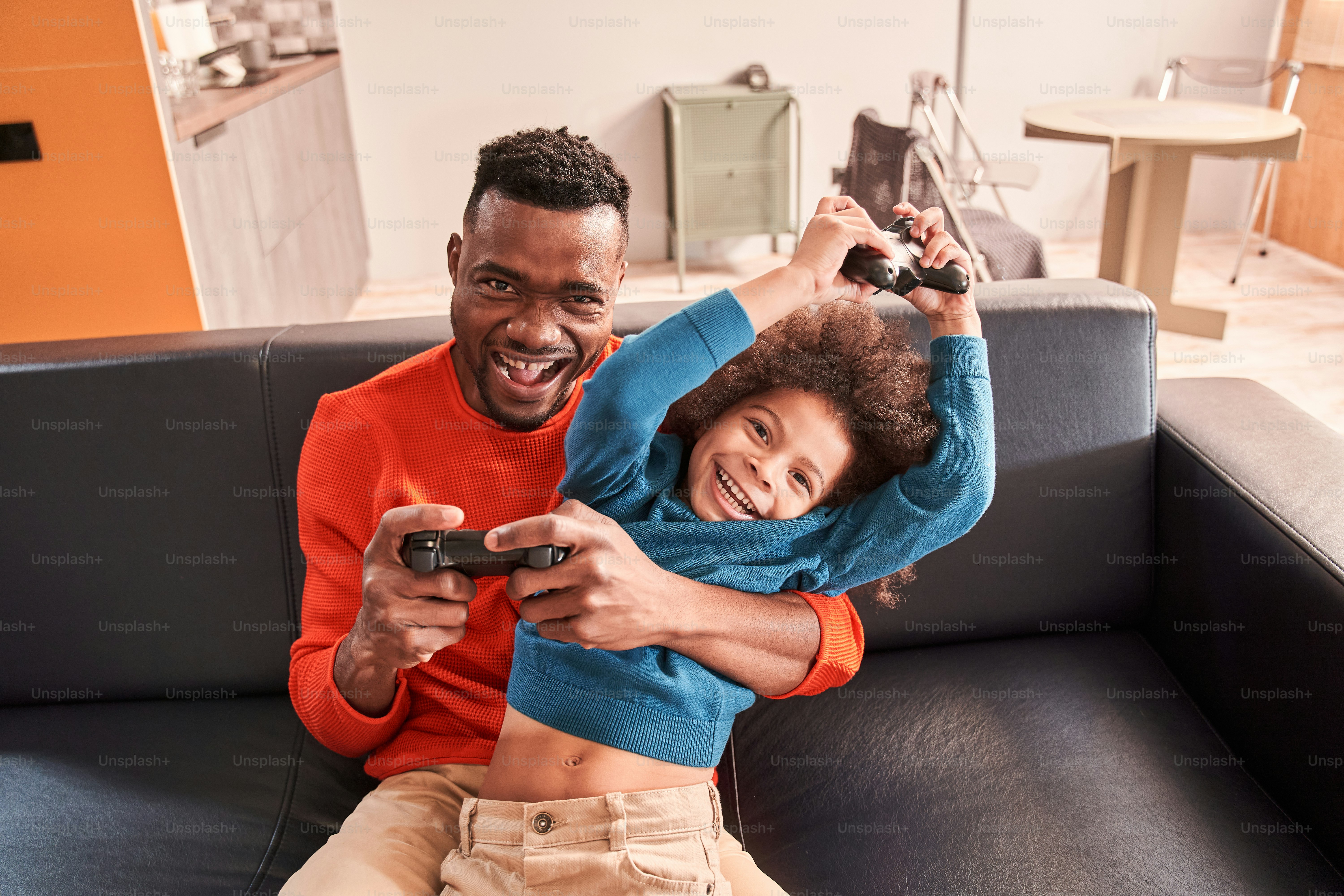 Smiling young bearded father and his little curly son wearing casual clothes relaxing on a couch, playing video games while toothy smiling at home. Family rejoicing with joysticks. Playing with family concept