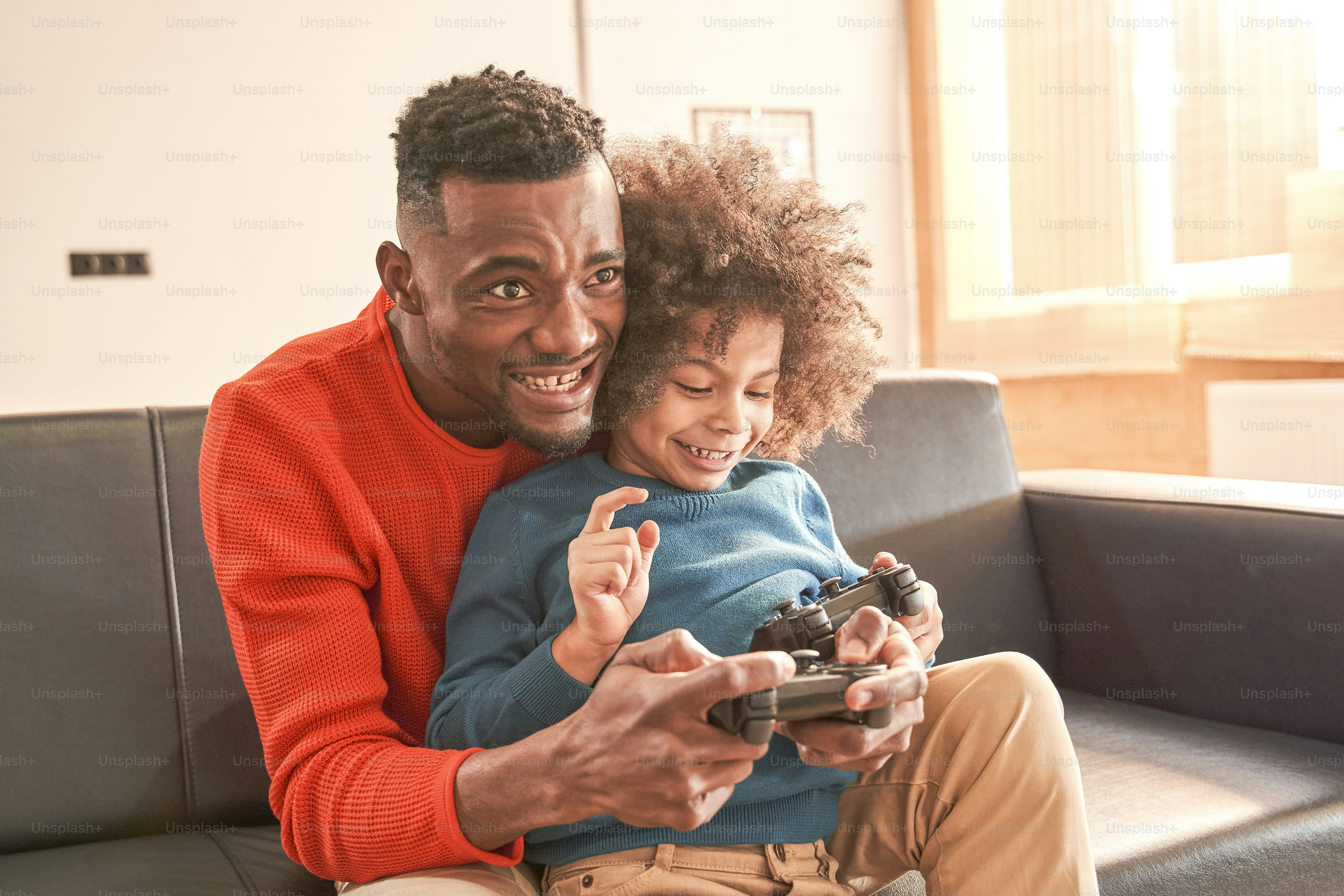 Waist up portrait view of the excited bearded father and his curly pretty son playing video game in living room while sitting at the sofa and holding joysticks. Games and entertainment concept