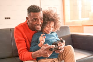 Vista de retrato de cintura hacia arriba del emocionado padre barbudo y su bonito hijo rizado jugando videojuegos en la sala de estar mientras están sentados en el sofá y sosteniendo joysticks. Concepto de juegos y entretenimiento