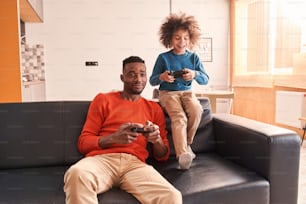 Happy bearded dad enjoying video game with his cute amazing curly son, while playing with joysticks at home. Childhood and father with son relationships concept. Stock photo