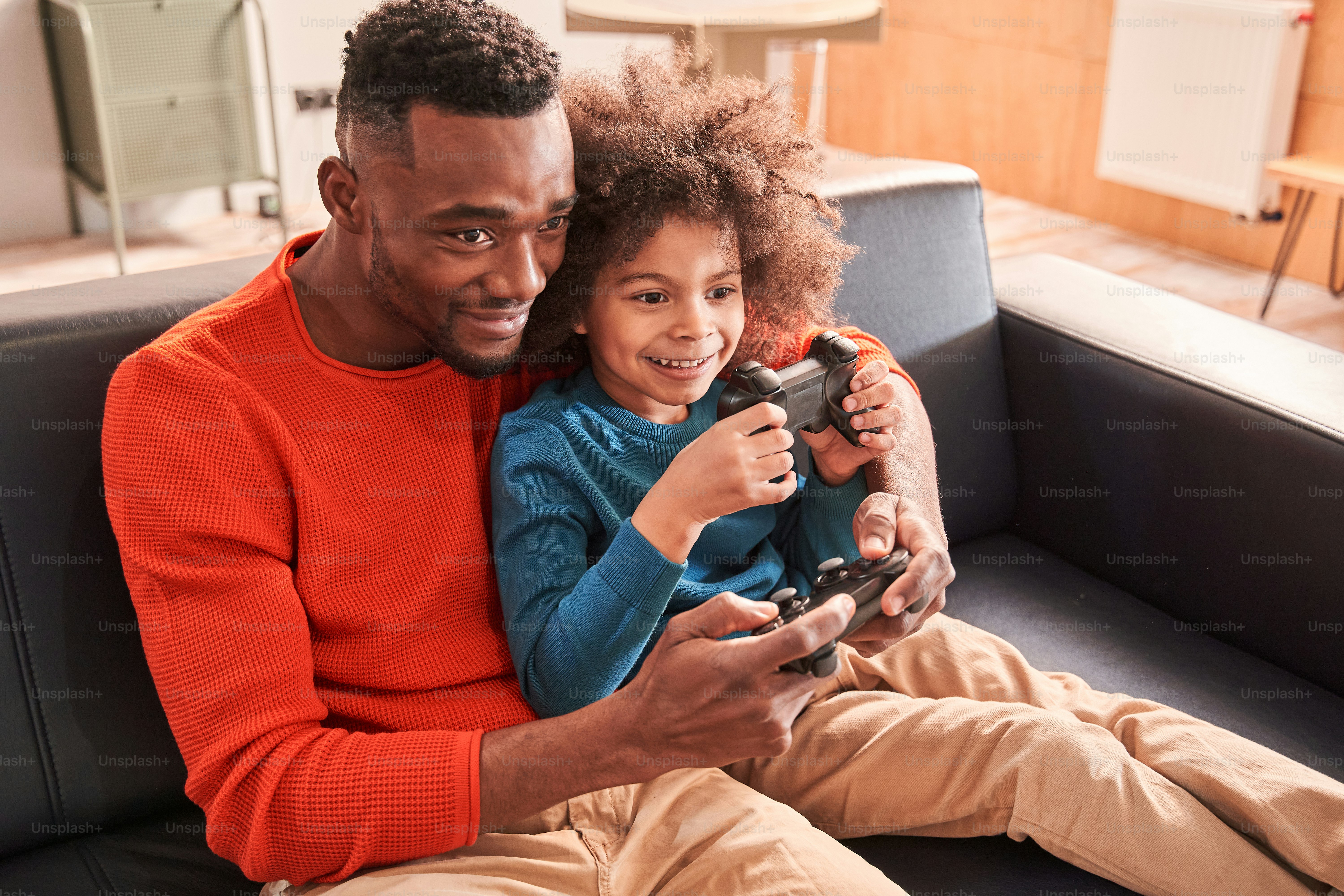 Waist up portrait view of the excited bearded father and his curly pretty son playing video game in living room while sitting at the sofa and holding joysticks. Games and entertainment concept
