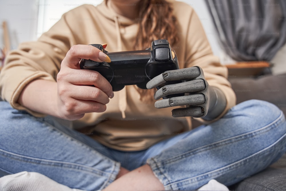 Cropped view of the caucasian young woman with artificial limb holding controller gamepad and playing video game at home while spending time at the sofa. Technology, gaming, entertainment and people concept