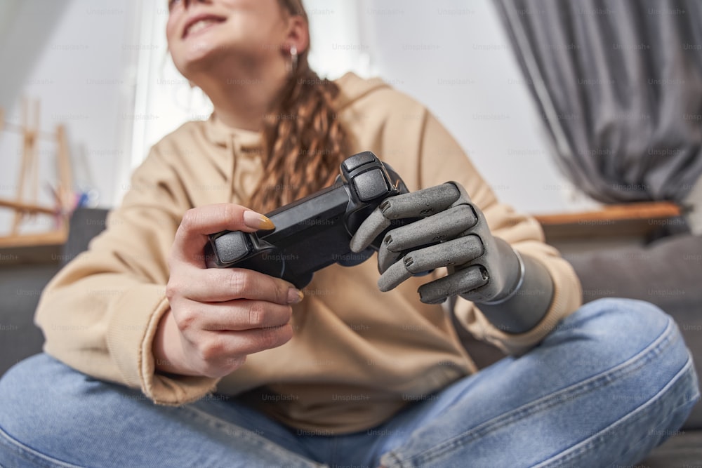 Vista de ángulo bajo de la mujer feliz involucrada con brazo de prótesis obteniendo nueva experiencia mientras usa gamepad en casa. Mujer jugando videojuegos con joystick mientras está sentada en el sofá