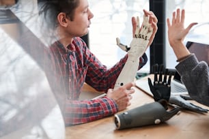 Compare it with my hand. Close up portrait of the male engineer looking attentively at the prosthesis hand and comparing it with real hand while working at the table with his colleague. Stock photo