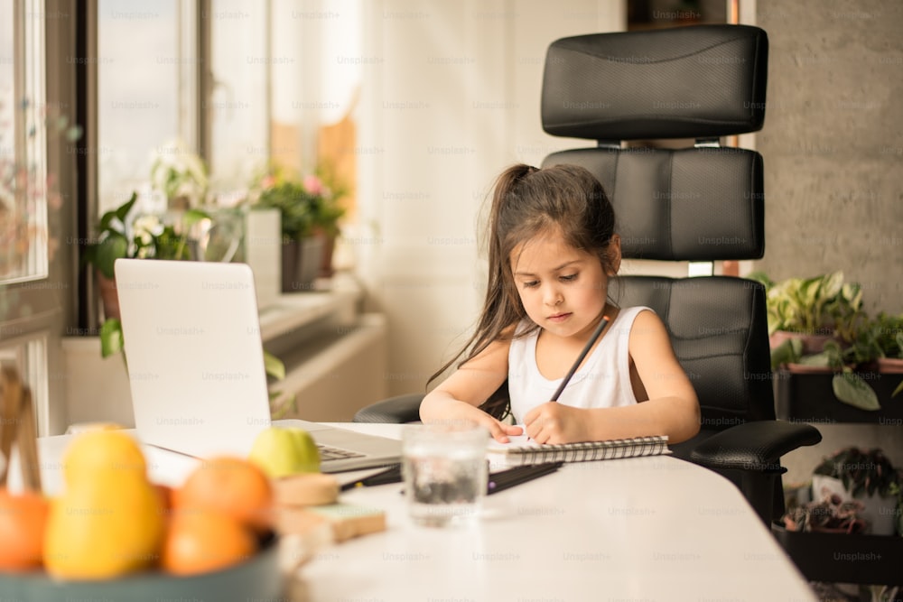 Pensive brunette girl puzzled about homework while studying at home isolation. Little kid writing at the notebook . Concept for quarantine and self isolation