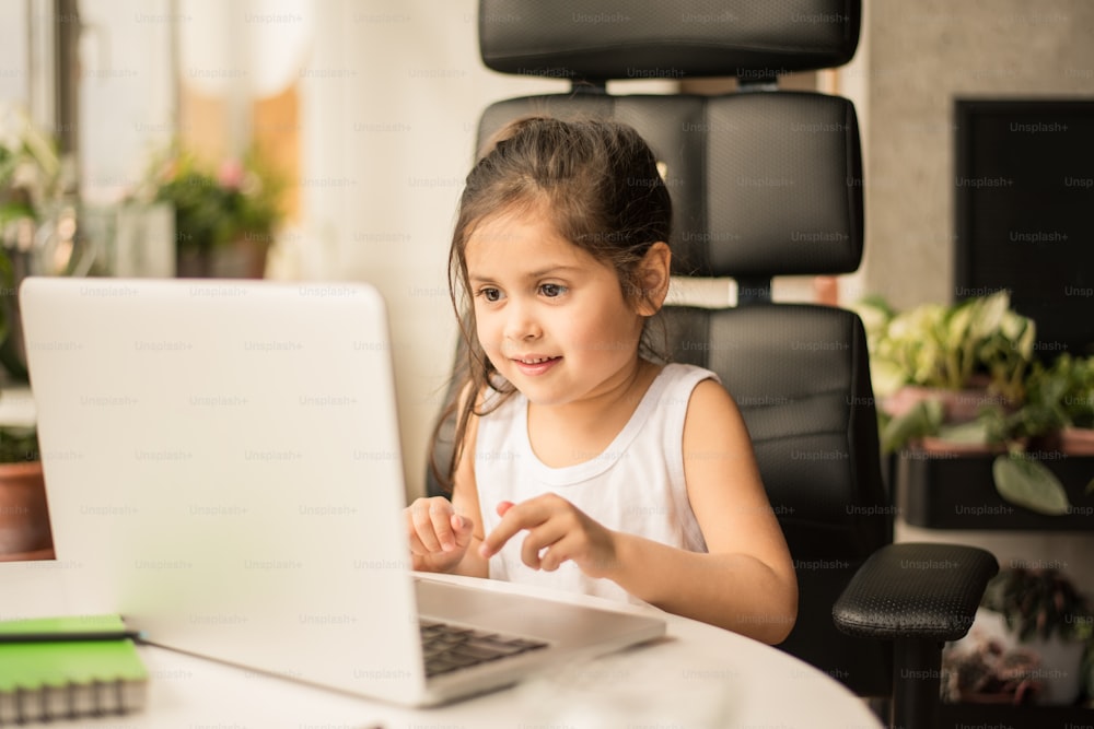 Pensive brunette girl puzzled about homework while studying at home isolation. Little kid looking at the laptop screen and smiling. Concept for quarantine and self isolation