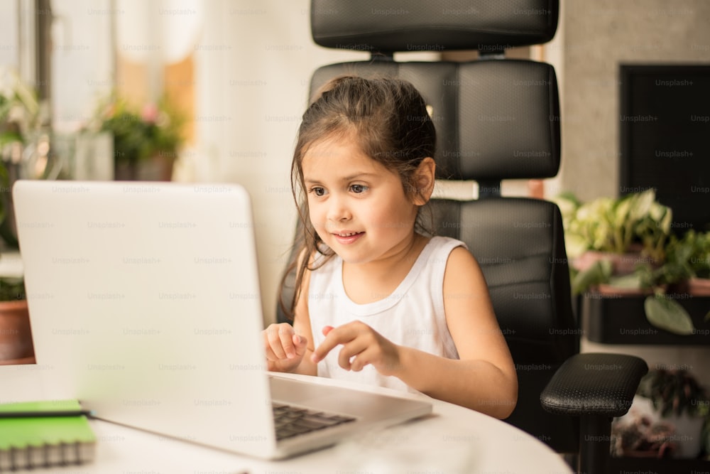物思いにふけるブルネットの女の子は、自宅で孤立して勉強しながら宿題に戸惑いました。ノートパソコンの画面を見て微笑む小さな子供。検疫と自己隔離のコンセプト