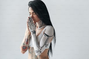 So tender. Horizontal portrait of a tattooed girl with cyborg body art at her hand posing with closed eyes and prayer gesture at the studio. Human and cyborg concept