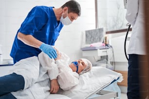 Full length view of the serious doctor talking with his patient woman laying at the hospital bed before the colonoscopy