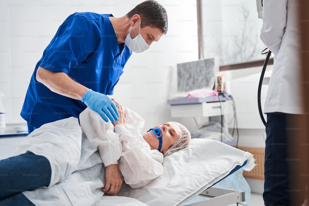 Visão completa do médico sério conversando com sua paciente deitada na cama do hospital antes da colonoscopia