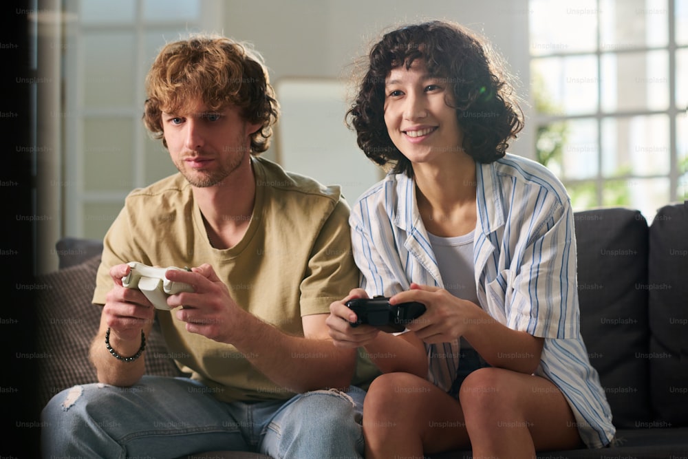 Happy young woman and her upset boyfriend in casual attire sitting on couch in living room at leisure and playing video game