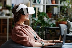 Side view of young female programmer in vr headset using laptop to control work with virtual stuff while sitting in openspace office